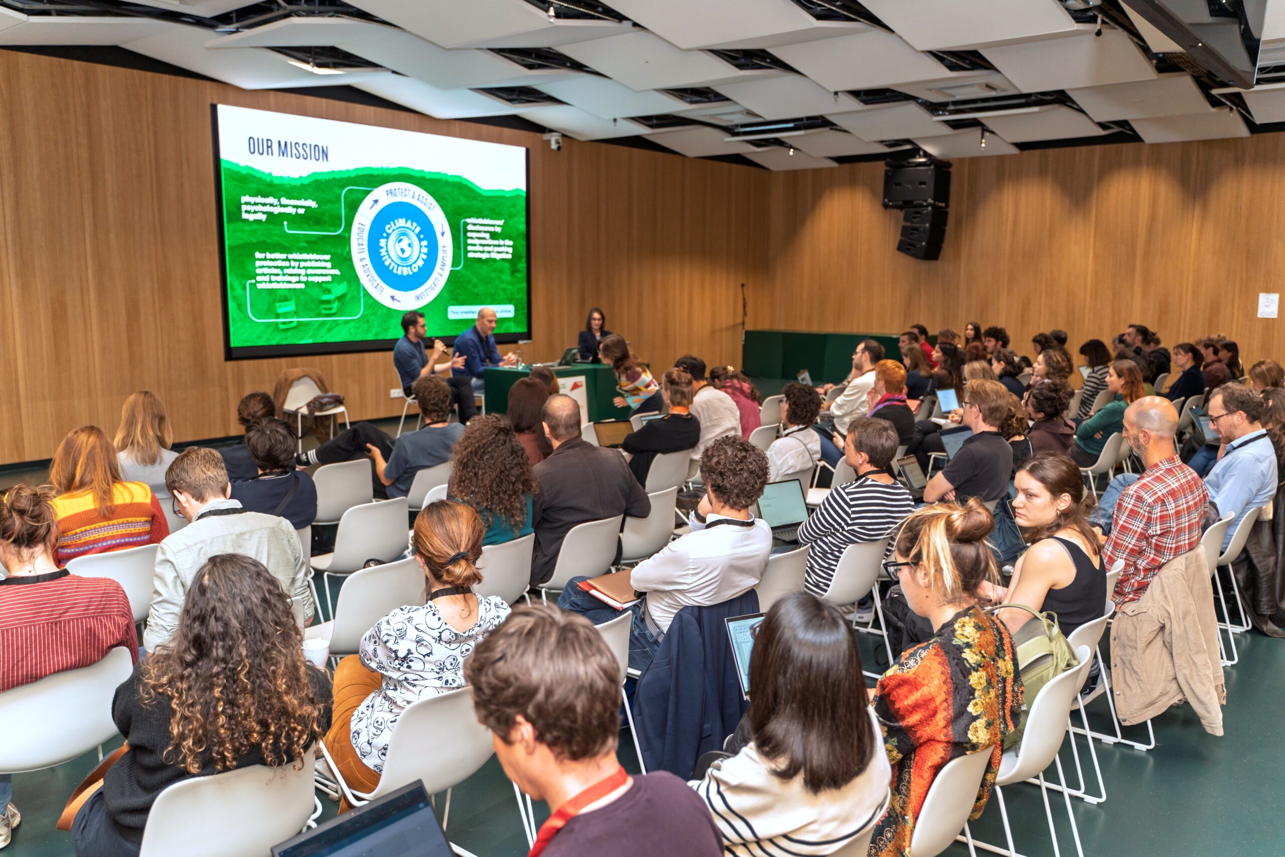A conference room full of people, sitting and looking at a panel of speakers. Behind the panel is a powerpoint about the mission of a climate whistleblower protection organisation.